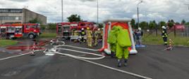 Strażacy w ubraniach gazoszczelnych ćwiczą na terenie zakładu. Samochody pożarnicze podają wodę do rozłożonej kabiny dekontaminacyjnej. 