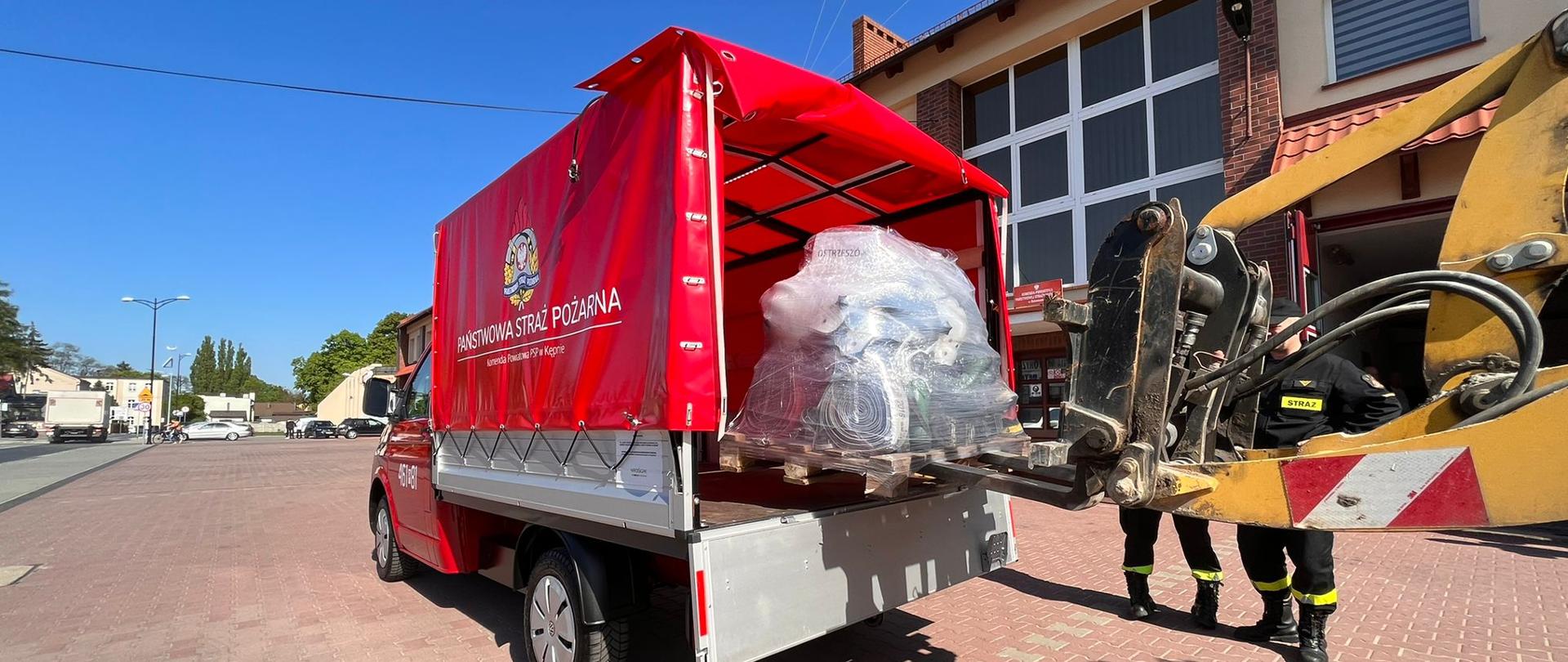 Ładowacz czołowy pakuje paletę na samochód dostawczy. Zdjęcie zrobione na placu manewrowym przed budynkiem komendy.