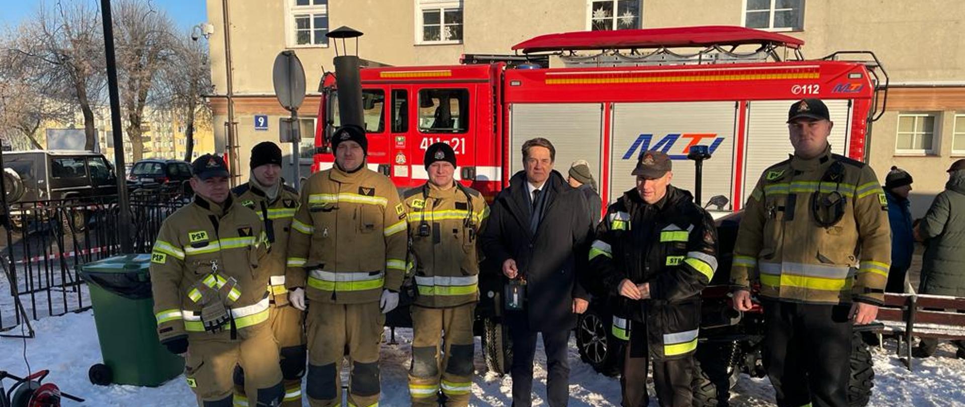 Zima, biało od śniegu. Na zdjęciu starosta braniewski stoi ze strażakami w jasnych mundurach. W tle budynek i samochód strażacki. Słonecznie.