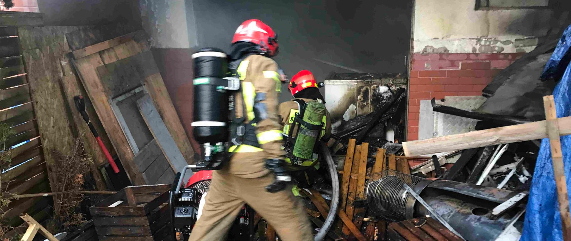 Zdjęcie przedstawia dwóch strażaków podających prąd wody do wnętrza garażu. Strażacy pracują w sprzęcie ochrony dróg oddechowych. 