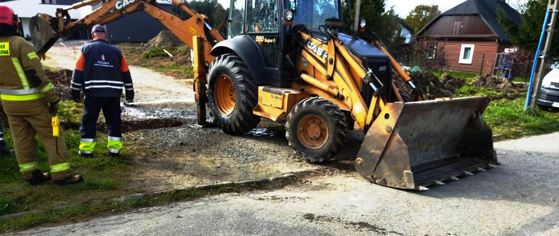 Na zdjęciu widać koparkę odkopującą dalszą część uszkodzonej rury gazowej. Przy koparce stoi pracownik Polskiej spółki gazownictwa oraz strażak w umundurowaniu bojowym z urządzeniem pomiarowym do gazów niebezpiecznych. W tle widać budynki.