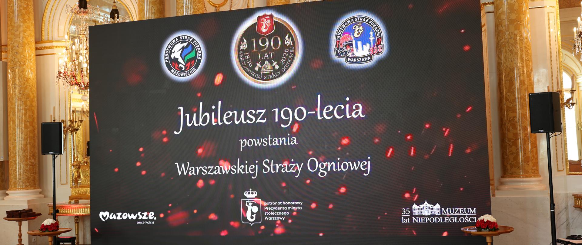 W złotej sali Zamku Królewskiego widoczny jest duży ekran. Na czarnym tle znajdują się logo Komendy Wojewódzkiej PSP w Warszawie, Komendy Miejskiej PSP m. st. Warszawy oraz logo 190-lecia Warszawskiej Straży Ogniowej, a także biały napis „Jubileusz 190-lecia powstania warszawskiej Straży Ogniowej”. 