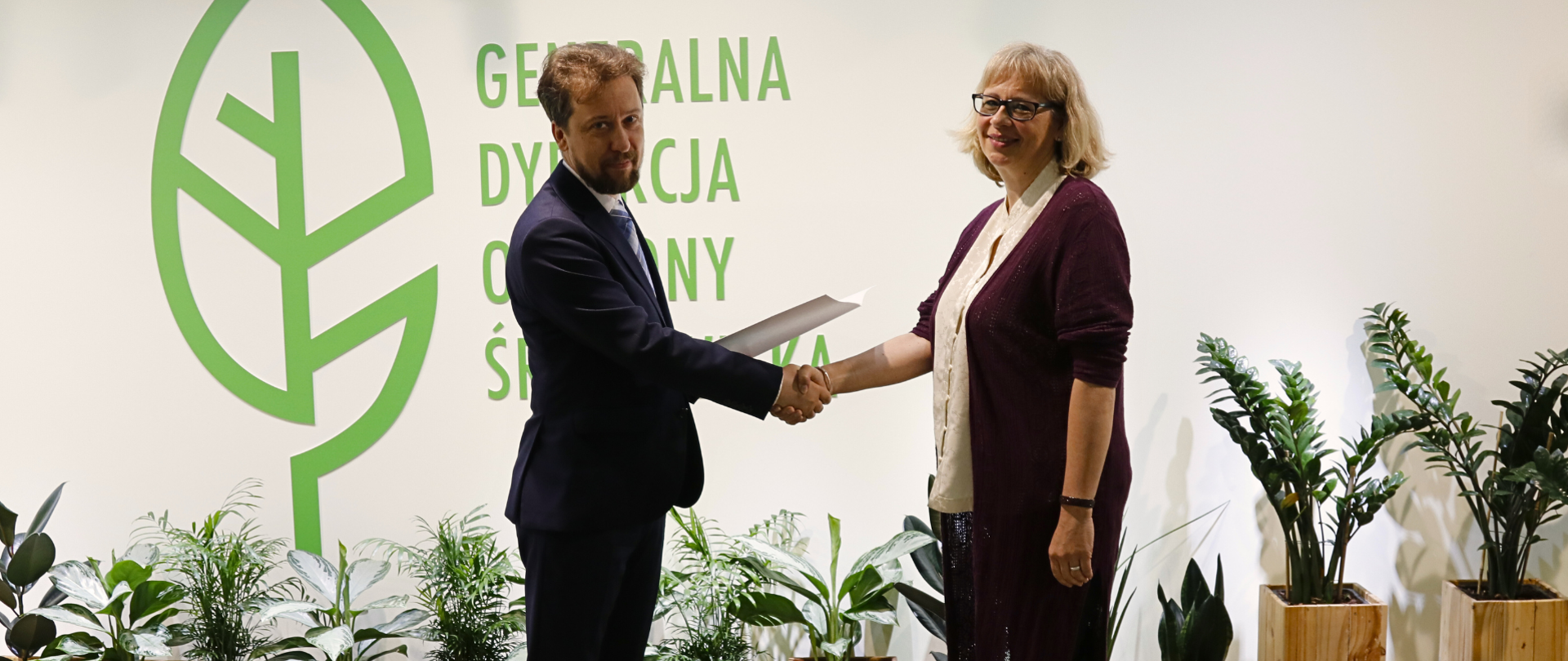 Na tle białej ściany z logotypem Generalnej Dyrekcji Ochrony Środowiska (zielony liść) stoi mężczyzna w garniturze i kobieta. Mężczyzna ściska dłoń kobiety. Mężczyzna trzyma w drugiej dłoni teczkę. 