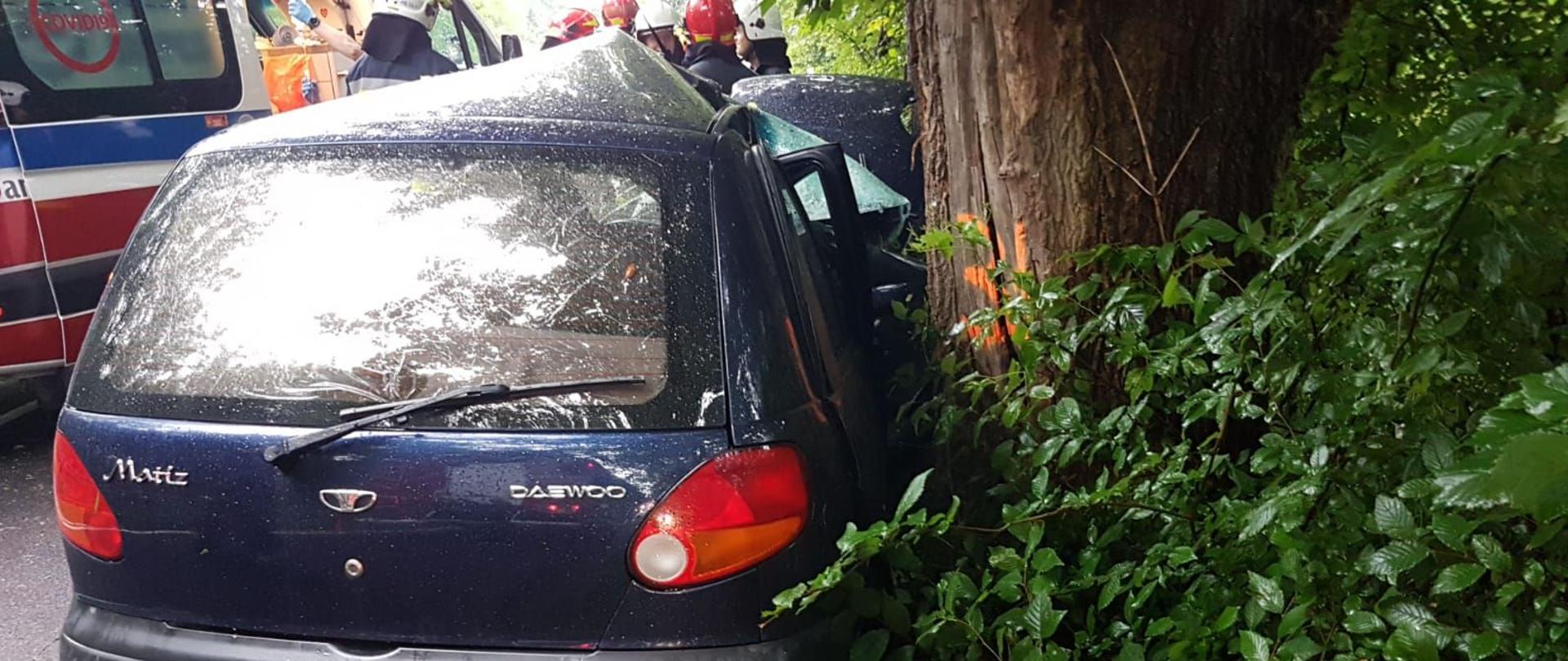 Samochód osobowy koloru granatowego stoi prawym bokiem do drzewa, cały bok jest wgnieciony. W tle z lewej strony karetka pogotowia.