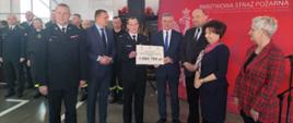 Zdjęcie przedstawia Wielkopolskiego Komendanta Wojewódzkiego PSP, Panią Minister oraz Pana Ministra wręczających promesę na zakup samochodu ratowniczo-gaśniczego. W tle stoją funkcjonariusze KP PSP w Gostyniu.