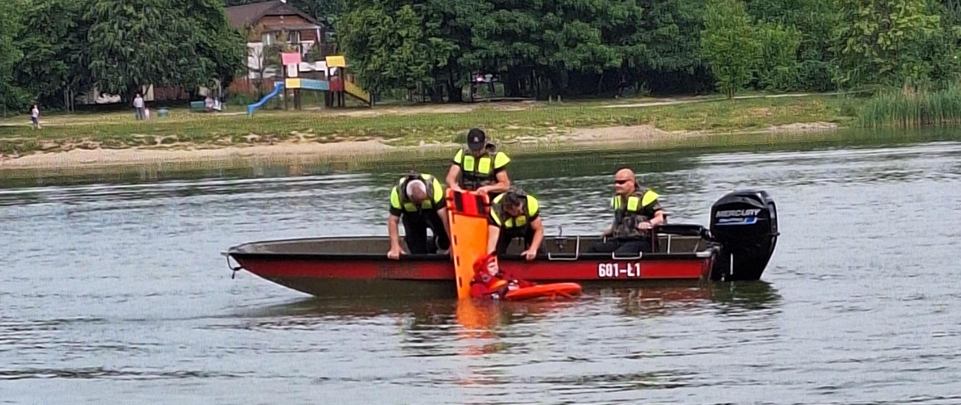 Zdjęcie wykonane z brzegu, na wodzie pływa łódź strażacka. W łodzi jest czterech ratowników, którzy wyciągają z wody strażaka w czerwonym skafandrze.