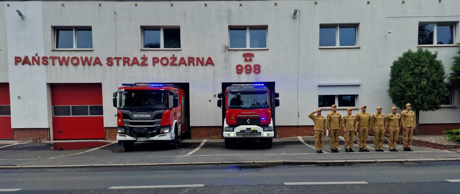 Budynek straży pożarnej z napisem „PAŃSTWOWA STRAŻ POŻARNA” i numerem alarmowym „998” na elewacji. Przed dwoma otwartymi garażami stoją dwa czerwone samochody ratowniczo-gaśnicze z włączonymi niebieskimi światłami ostrzegawczymi. Po prawej stronie pojazdów stoi w szeregu osiem osób w umundurowaniu, z których jedna wykonuje salut. Na pierwszym planie widoczna jest jezdnia, a po prawej stronie budynku rośnie duże drzewo. Niebo jest pochmurne.
