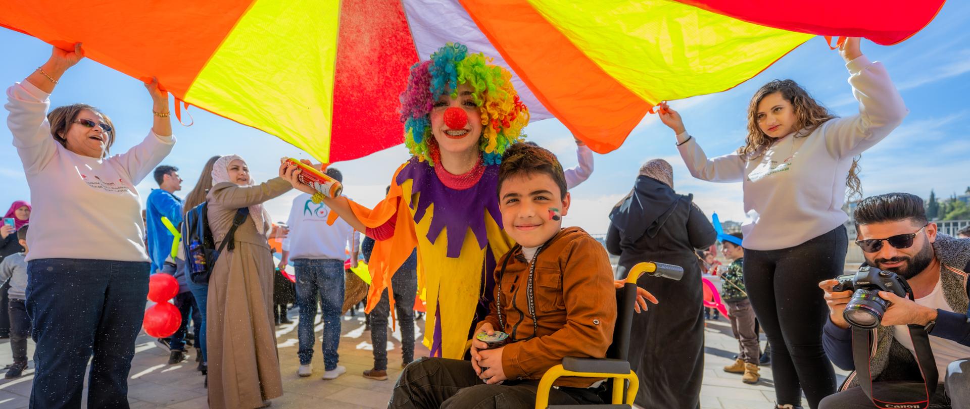 Uśmiechnięty chłopiec na wózku inwalidzkim z namalowaną flagą Palestyny na policzku. Obok niech uśmiechnięty kolorowy klałn. Wokół nich śmiejący się ludzie. 
