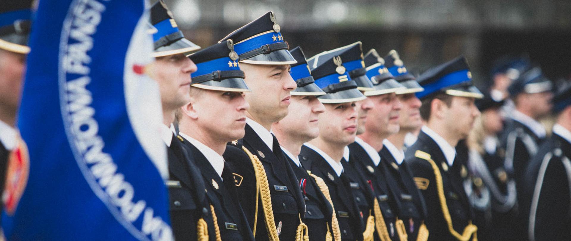 Zdjęcie przedstawia szereg funkcjonariuszy Państwowej Straży Pożarnej w mundurach galowych, stojących w równej formacji podczas oficjalnej uroczystości. Strażacy noszą czapki rogatywki z niebieskimi otokami oraz ozdobne złote sznury galowe