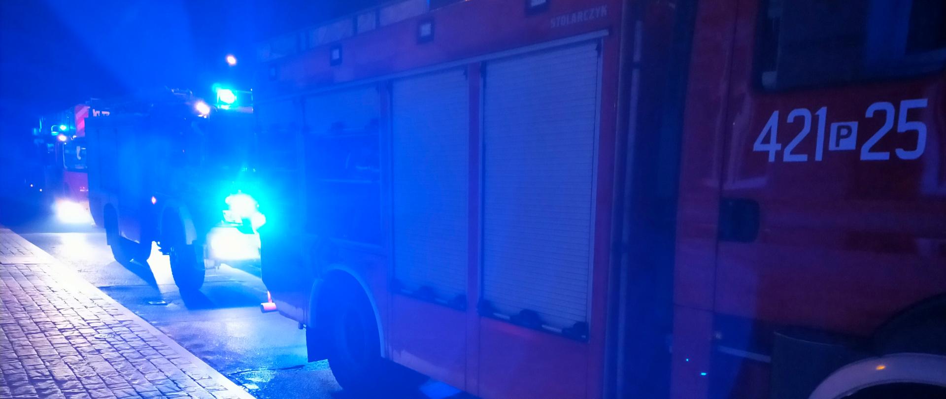 Zdjęcie zrobione w porze nocnej. Na zdjęciu widać trzy samochody strażackie stojące na ulicy jeden za drugim. Mają one włączone sygnały świetlne