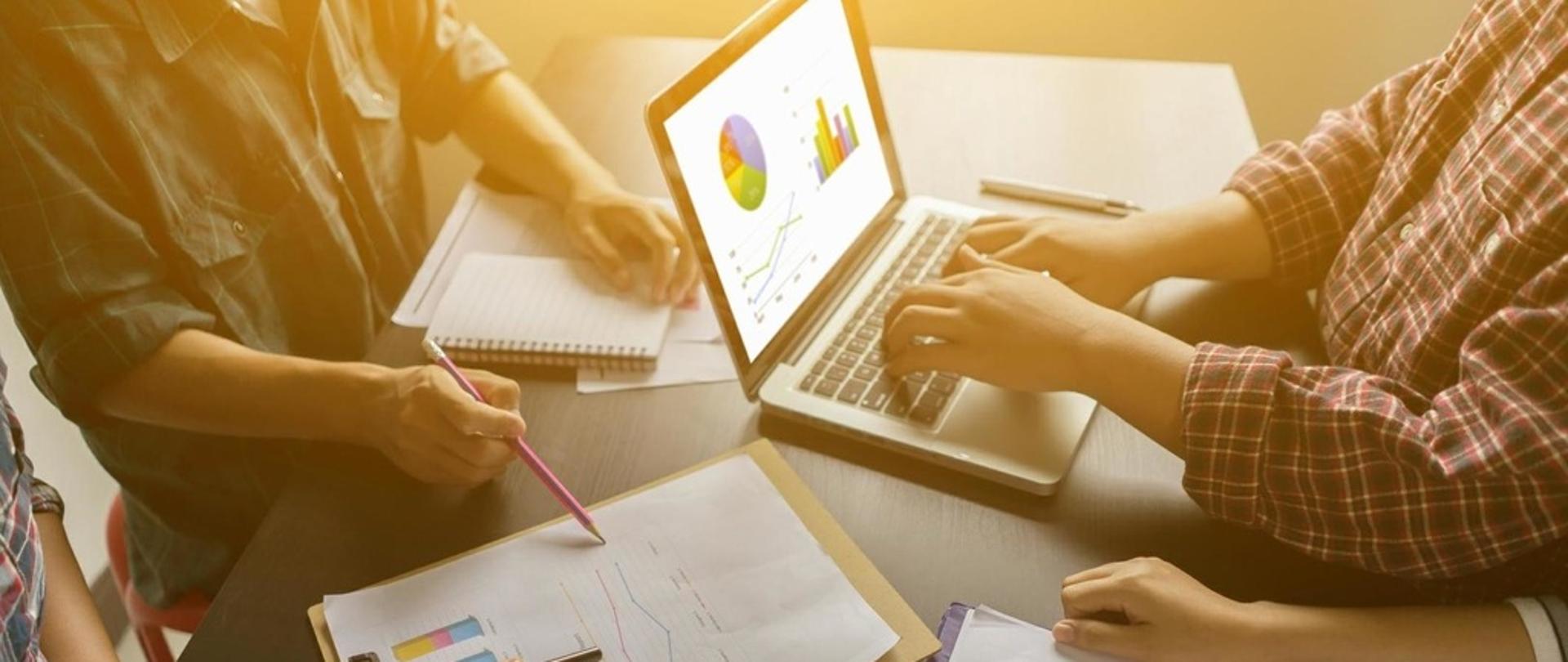 Cztery osoby siedzą przy stole. Na blacie stoi otwarty laptop oraz leżą dokumenty.