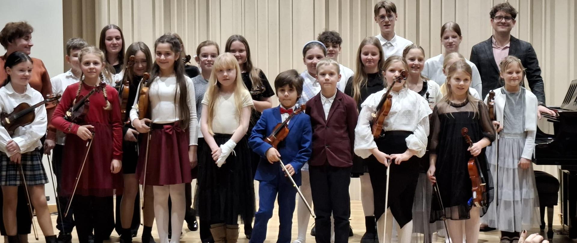 Grupa młodych uczniów występujących na koncercie klasy skrzypiec i fortepianu stoi razem na scenie, trzymając skrzypce i smyczki. W tle widoczni są również pedagodzy. Dzieci ubrane są odświętnie, uśmiechają się i pozują do wspólnej fotografii po zakończonym występie.