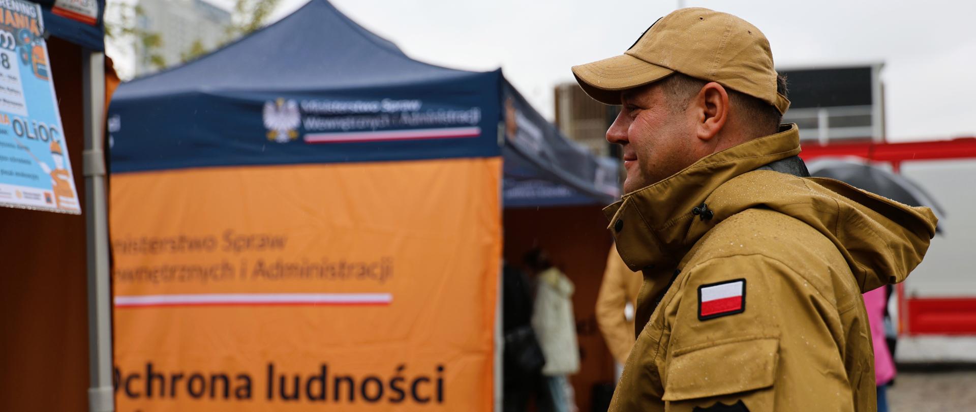 Na zdjęciu widoczny jest Mazowiecki Komendant Wojewódzki PSP nadbryg. Artur Gonera, ubrany w piaskowy mundur służbowy i czapkę z daszkiem. Został uchwycony z profilu podczas przechodzenia obok namiotu informacyjnego. W tle znajduje się granatowo-pomarańczowy namiot z widocznym napisem „Ochrona ludności i obrona cywilna”, a po lewej stronie częściowo widoczny jest czerwony samochód pożarniczy. Pogoda jest pochmurna, ale bez intensywnych opadów. Zdjęcie sprawia wrażenie spokojnego i pokazuje zaangażowanie w działania edukacyjne dotyczące bezpieczeństwa mieszkańców.
