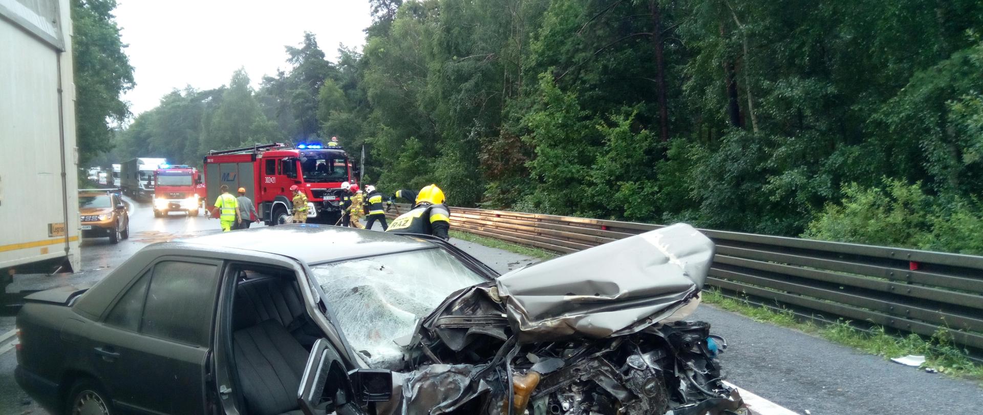 Zdjęcie przedstawia rozbitego mercedesa - pogięty cały przód. Droga jest mokra - widać rozsypany sorbent. W tle stoi samochód Straży Pożarnej.