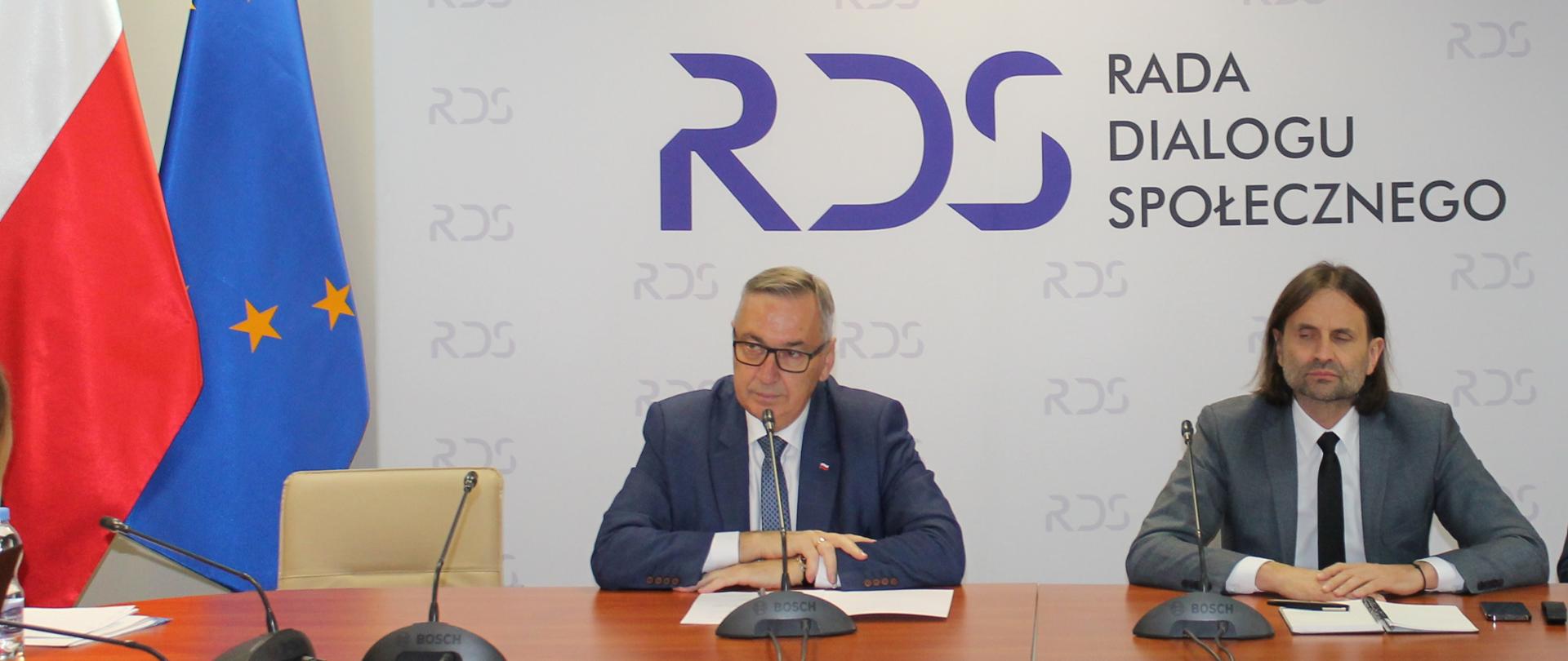 Zdjęcie przedstawia stół prezydialny za którym siedzą: Stanisław Szwed - Sekretarz Stanu w Ministerstwie Rodziny i Polityki Społecznej, Piotr Bromber - Podsekretarz Stanu w Ministerstwie Zdrowia, Współprzewodniczący Zespołu ze strony rządowej. W tle siedzących znajduje się baner z napisem Centrum Partnerstwa Społecznego Dialog im. Andrzeja Bączkowskiego. Po lewej stronie zdjęcia stoją dwie flagi: Polska i UE. Nad banerem widnieje Godło Polski.