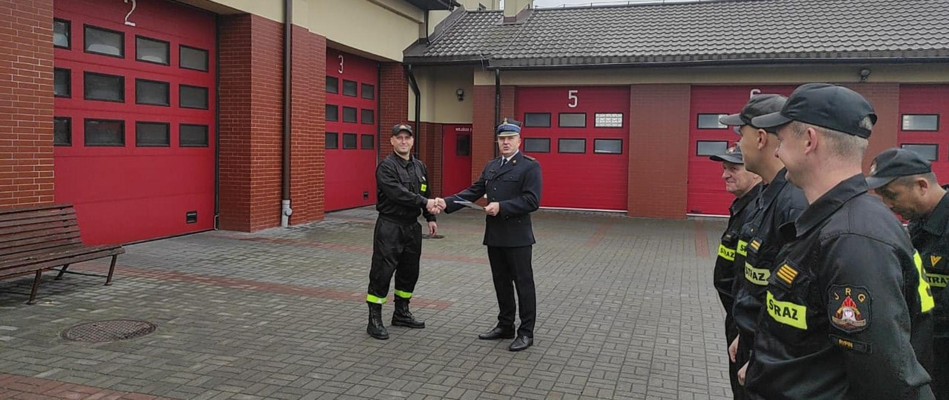 Starszy brygadier Grzegorz Łydkowski wręcza list gratulacyjny starszemu ogniomistrzowi Radosławowi Fodrowskiemu. Po prawej stronie zdjęcia stoją strażacy w czarnych ubraniach koszarowych na tle czerwonych bram garażowych. Strażacy stoją na szarym polbruku.