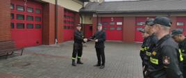 Starszy brygadier Grzegorz Łydkowski wręcza list gratulacyjny starszemu ogniomistrzowi Radosławowi Fodrowskiemu. Po prawej stronie zdjęcia stoją strażacy w czarnych ubraniach koszarowych na tle czerwonych bram garażowych. Strażacy stoją na szarym polbruku.