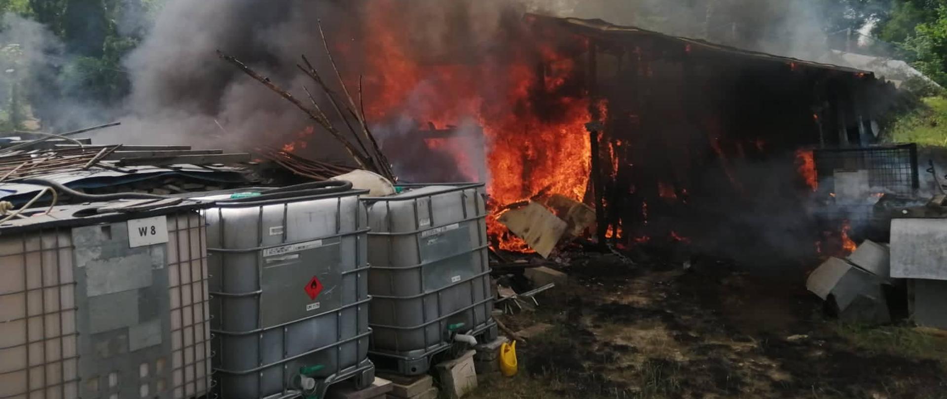 Na zdjęciu widoczny cały budynek gospodarczy w płomieniach. Przed budynkiem stoją zbiorniki z wodą.
