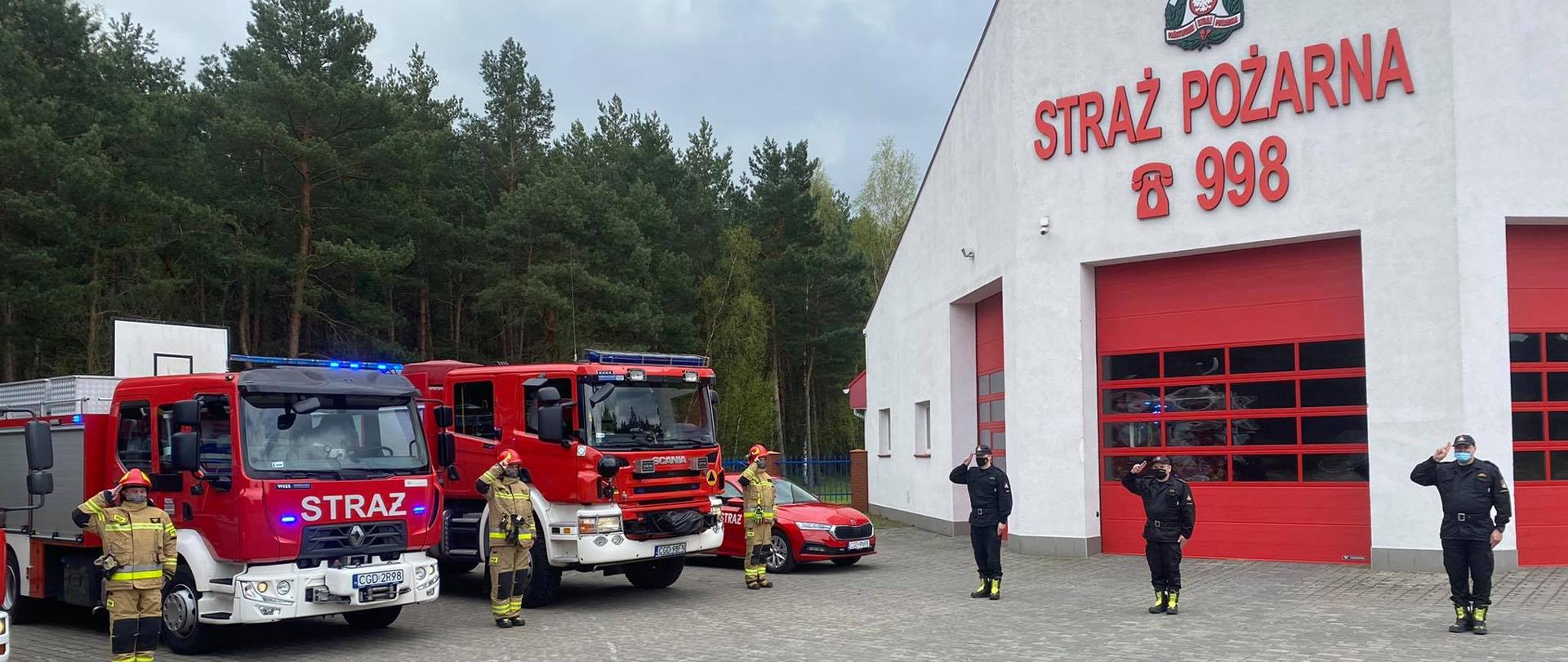 Na zdjęciu widać po lewej stronie widać 2 wozy strażackie oraz samochód operacyjny lekki , obok samochodów stoją strażacy w ubraniach specjalnych, po prawej stronie stoi 3 strażaków w mundurach dowódczo-sztabowych, w tle widać budynek komendy .
