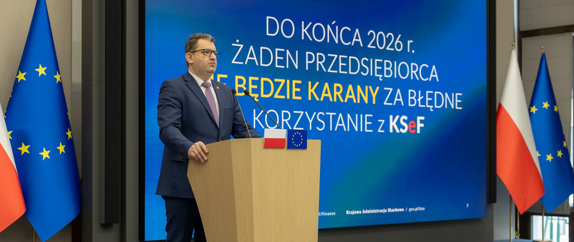Marcin Łoboda, Sekretarz Stanu w Ministerstwie Finansów, Szef Krajowej Administracji Skarbowej, stoi za mównicą, w tle ekran z prezentacją o KSeF oraz flagi Polski i UE