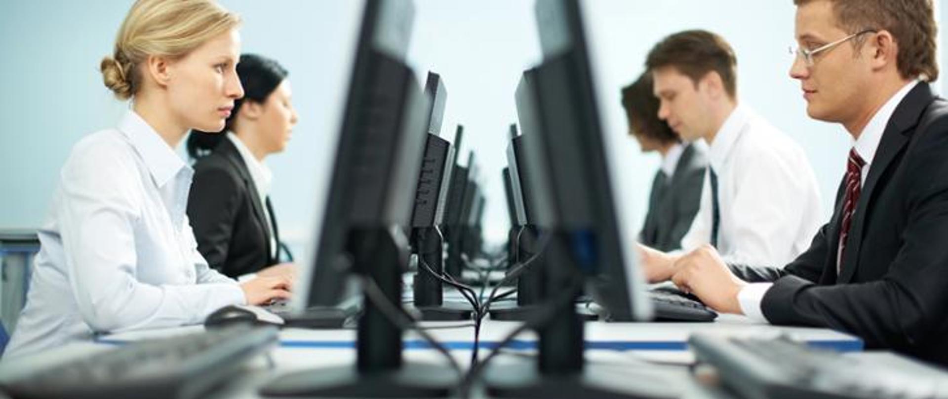 Na pierwszym planie dwoje pracowników biurowych zasiadający przy komputerach. W tle kolejni pracownicy biurowi.