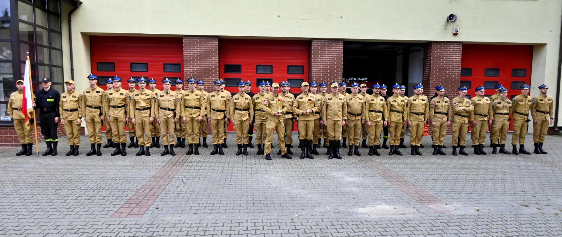 Na zdjęciu stoją w dwuszeregu strażacy PSP w piaskowych mundurach służbowych, a także w charakterystycznych dla kompanii reprezentacyjnej butach, paskach oraz czapkach rogatywkach. W tle strażacy PSP w piaskowych mundurach służbowych stoją w szeregu. Przed nimi widoczna kostka brukowa, a za nimi budynek i garaże strażackie. 