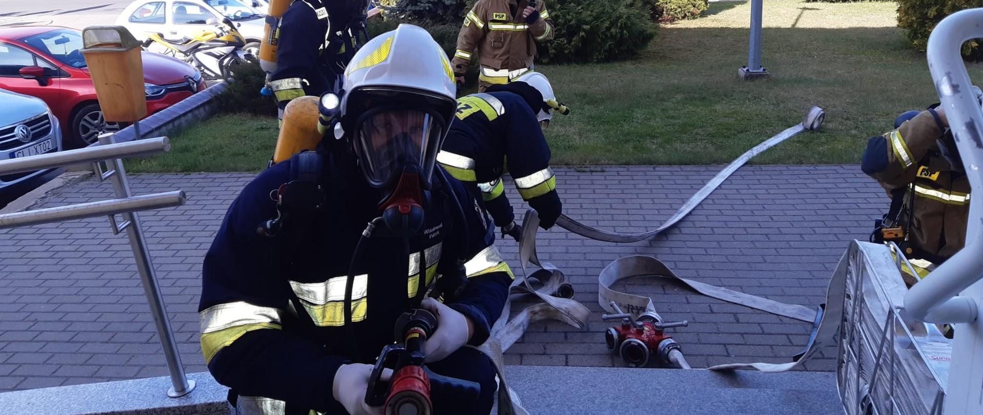 Zdjęcie przedstawia strażaków w nomeksach, hełmach i aparatach powietrznych. Strażacy mają linię wężową i przygotowują się do wejścia do budynku.