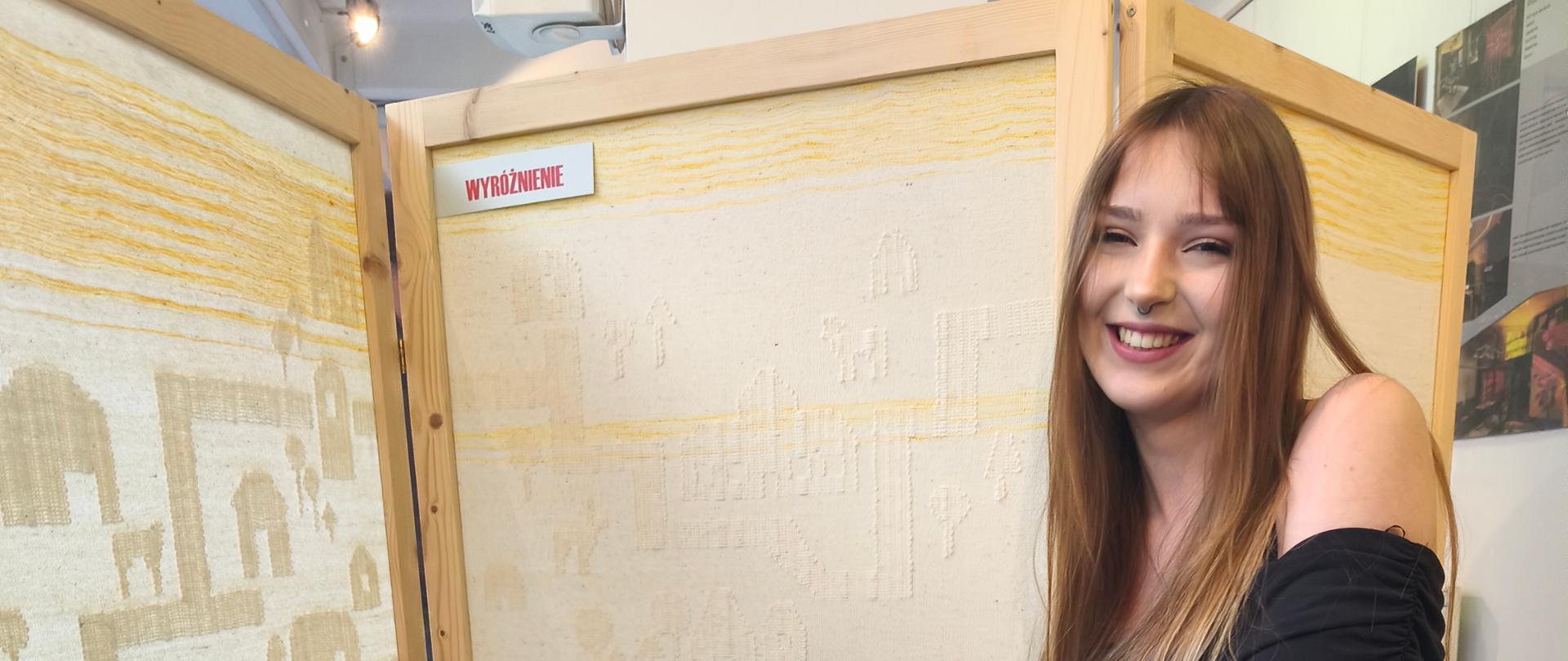 Dziewczyna ubrana w czarną sukienkę siedzi bokiem na krześle na tle parawanu z wypełnieniem w technice tkackiej