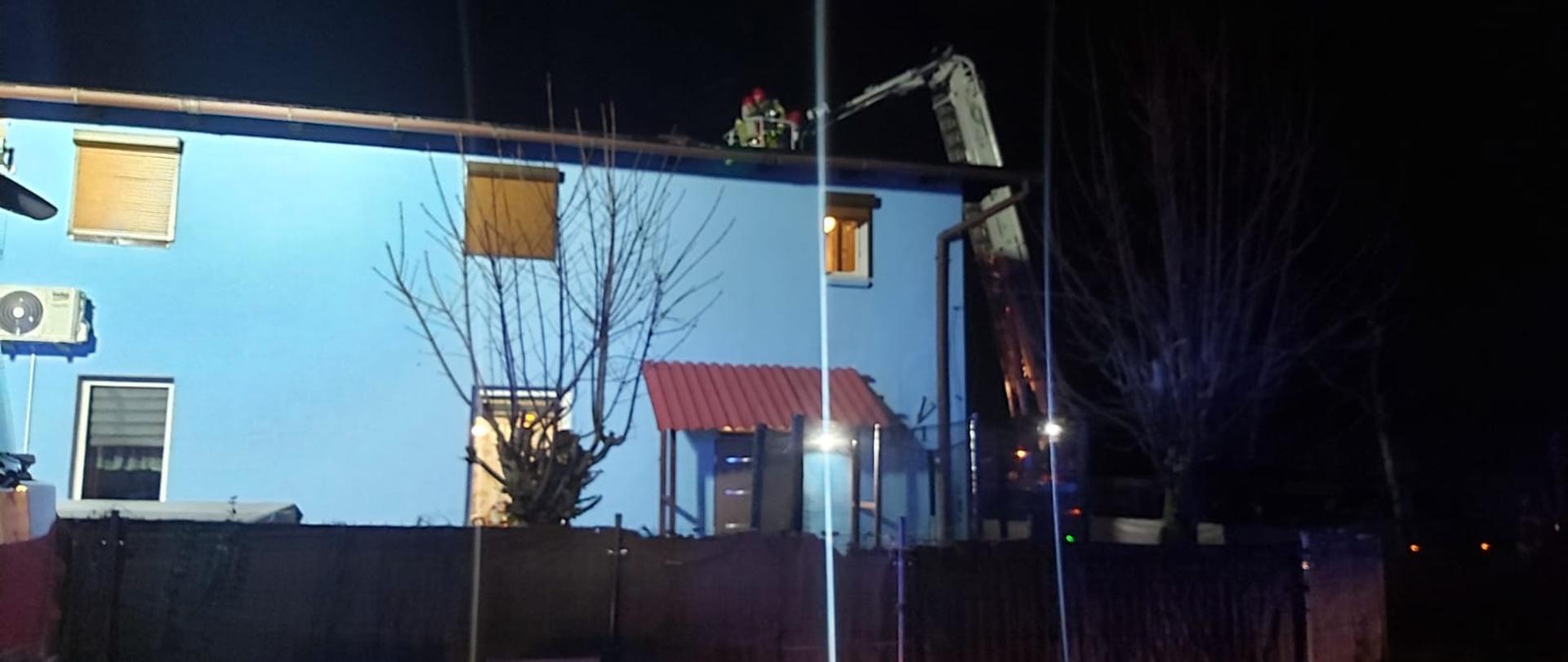 NA zdjęciu budynek z boku budynku w koszu podnośnika strażacy wykonujący czynności służbowe - gaszą pożar sadzy w kominie