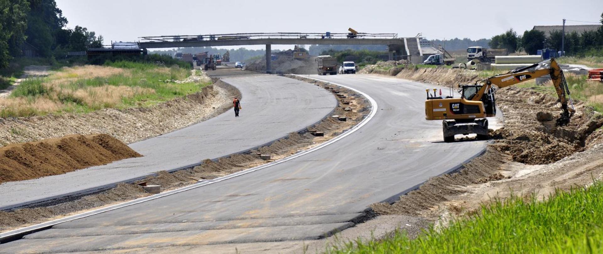 Budowa drogi ekspresowej – widoczny odcinek nowej jezdni w trakcie prac, po prawej stronie koparka marki CAT wykonuje roboty ziemne, w tle kilka pojazdów budowlanych i most w budowie nad trasą, po lewej nasypy ziemne i pas zieleni.