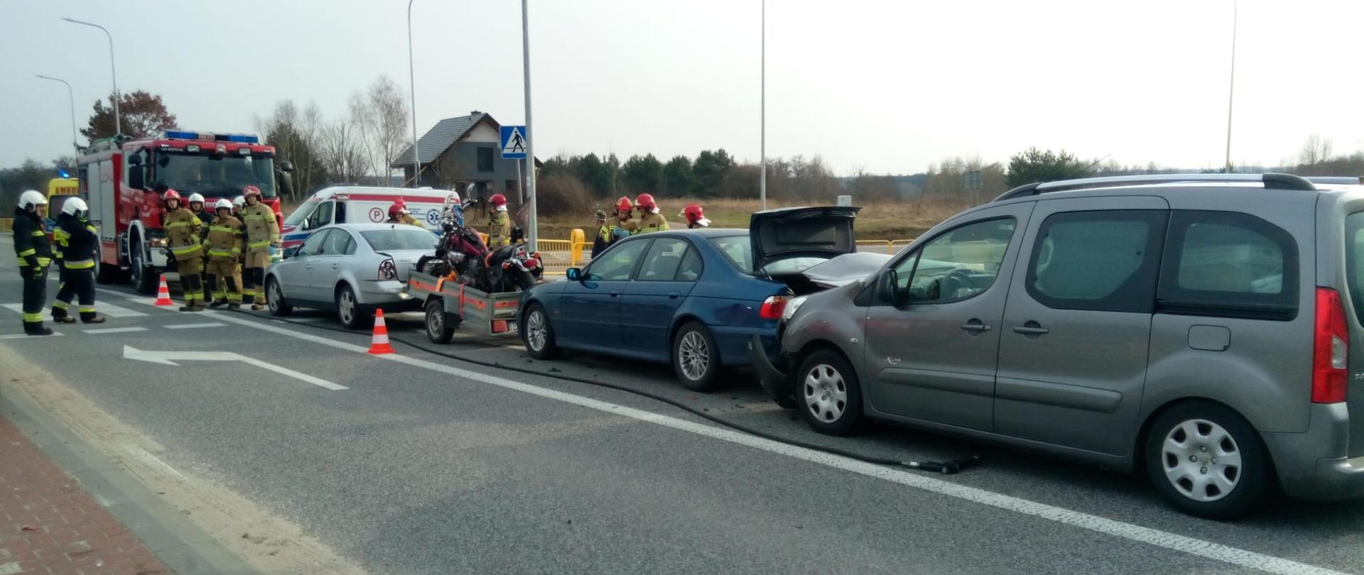 Zdjęcie przedstawia wszystkie biorące udział w wypadku samochody stojące na pasie drogowym. W głębi widać pojazdy służb ratowniczych oraz stojących strażaków. 