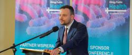 Uśmiechnięty mężczyzna w garniturze nachyla się nad mównicą i mówi do mikrofonu na stojaku, za nim przed żółtą ścianą dwa rozwinięte niebiesko-różowe banery z napisami Konferencja „Elbląg PUBLIC HEALTH”.