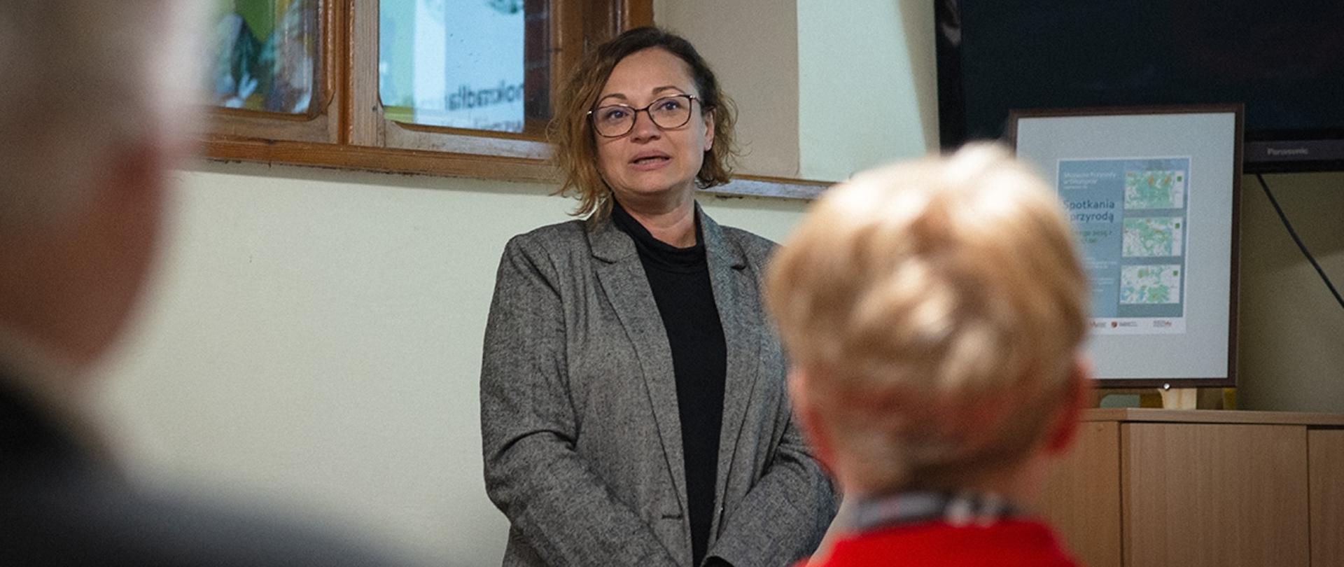 Na pierwszym planie grupa siedzących ludzi a sali, na drugim planie Pani Aleksandra Krzysztoń-Rzodkiewicz - p.o. Zastępcy Regionalnego Dyrektora Ochrony Środowiska w Olsztynie, Regionalnego Konserwatora Przyrody siedząca przy komputerze opowiadająca o roli obszarów wodno-błotnych.