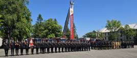 Kompania PSP z pocztem sztandarowym maszeruje w zwartej formacji. W tle stoją strażacy w ubraniach specjalnych oraz liczne wozy bojowe. Nad placem rozpościera się niebieskie niebo.

