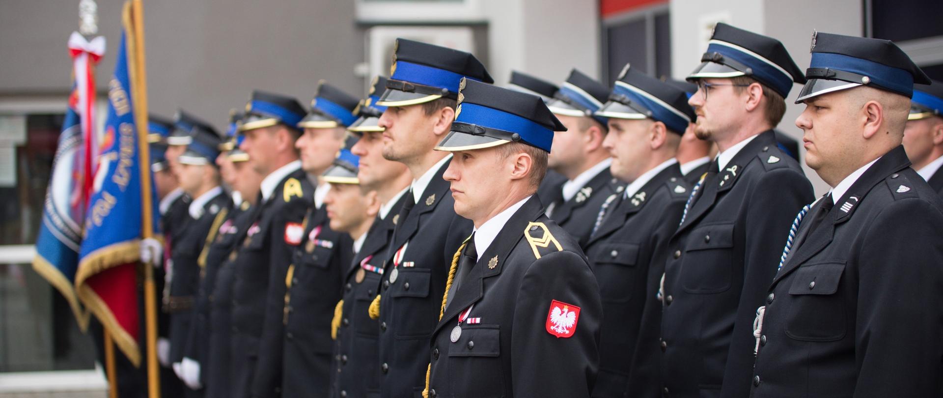 strażacy PSP i OSP w mundurach galowych, stojący w szeregach w oddali dwa poczty sztandarowe - obraz rozmazany