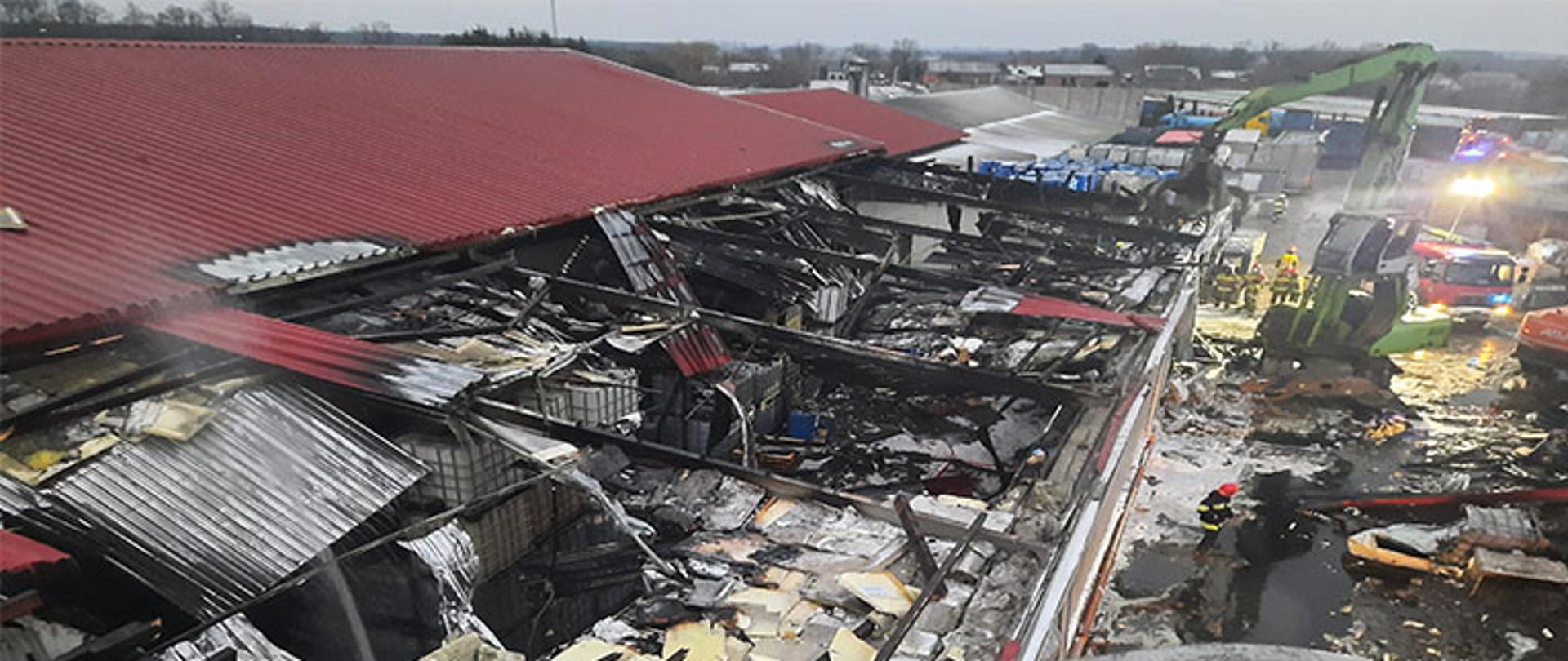 pogorzelisko i zerwany dach w hali produkcyjnej widoczne pojemniki z chemikaliami częściowe pokrycie hali czerwoną blachą falistą duża ilość materiałów izolacyjnych rozrzucona dookoła, koparka przeładunkowa w tle dwa pojazdy straży pożarnej woda z pianą na terenie placu