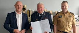 Komendant Powiatowy PSP w piaskowym mundurze stoi w rzędzie razem z druhem OSP i mężczyzną w garniturze. Mężczyzna znajdujący się pośrodku trzyma białą teczkę