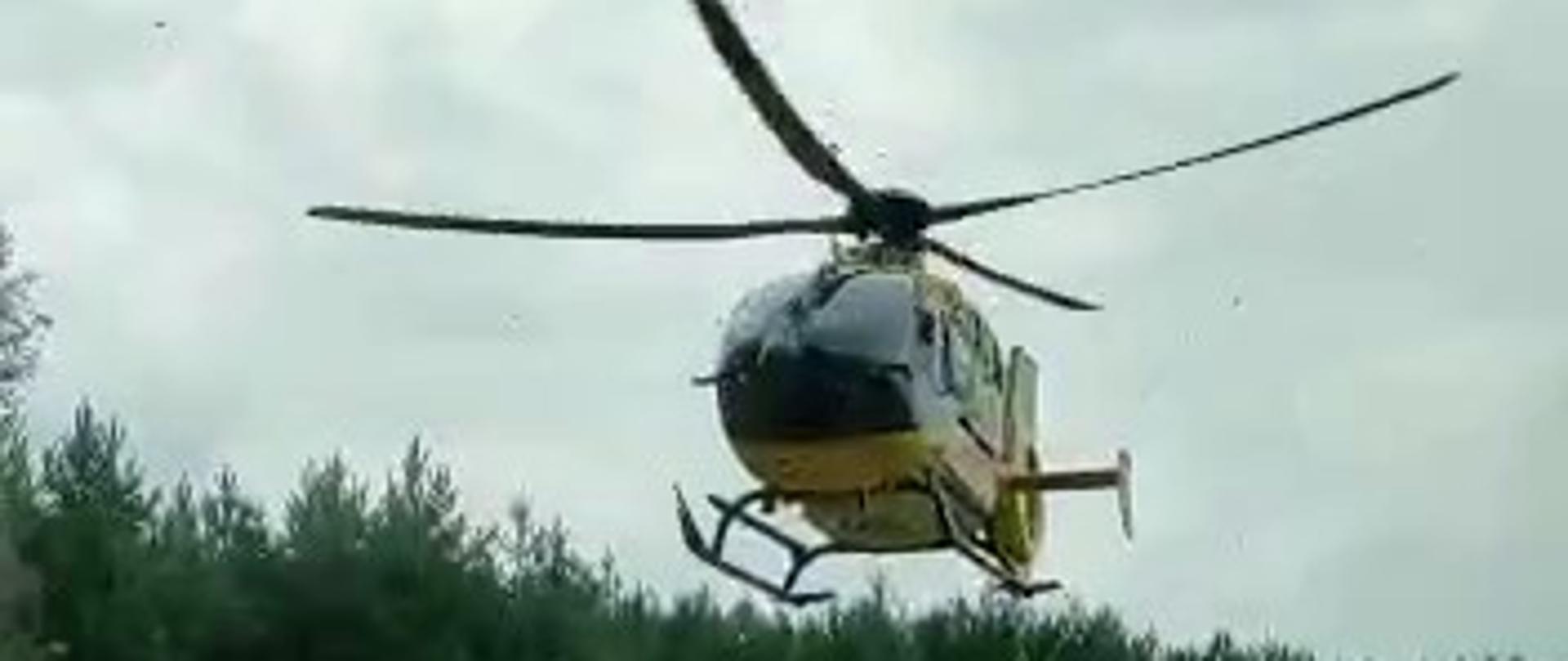 Zdjęcie przedstawia lądujący śmigłowiec Lotniczego Pogotowia Ratunkowego w pobliżu ulicy Cmentarnej w Kielcach w celu udzielenia pomocy poszkodowanemu w wypadku na motocyklu.
