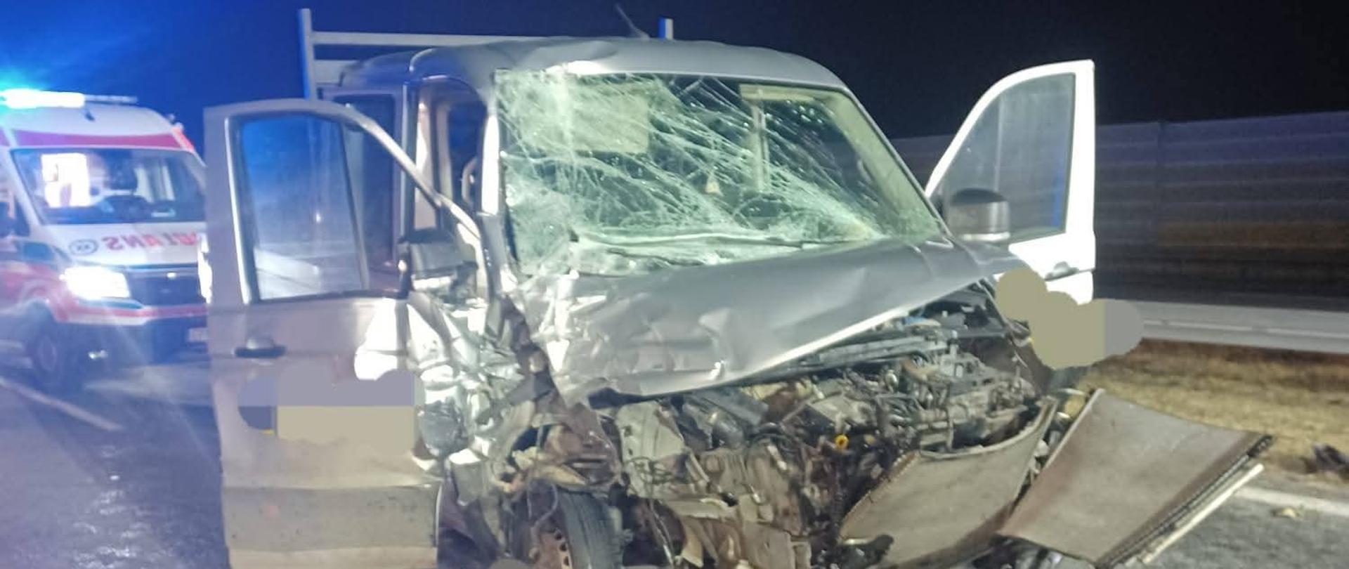 Na zdjęciu widać uszkodzony biały samochód dostawczy stojący na jezdni. Przód pojazdu jest całkowicie zgnieciony – maska jest wgnieciona, zderzak oderwany i leży na drodze, elementy silnika są odsłonięte. Przednia szyba jest mocno popękana. Przednie drzwi są otwarte. Na jezdni widać rozlane płyny eksploatacyjne. W tle, po lewej stronie, stoi karetka pogotowia z włączonymi niebieskimi światłami sygnalizacyjnymi. Po prawej stronie znajduje się bariera ochronna i ekran dźwiękochłonny, co sugeruje, że zdarzenie miało miejsce na autostradzie. Sytuacja rozgrywa się w nocy.