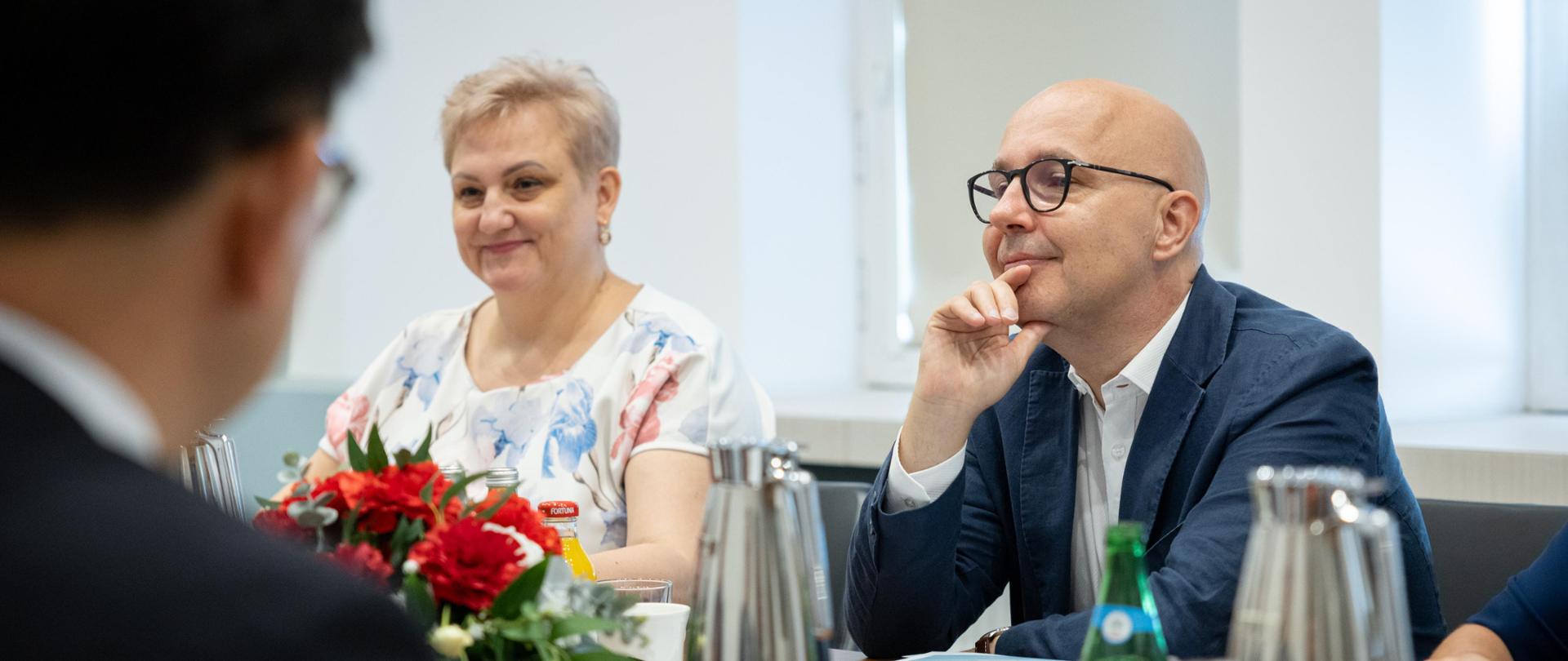 Przy stole siedzi kobieta i mężczyzna. Na stole widać dzbanki, kwiaty i butelki. 