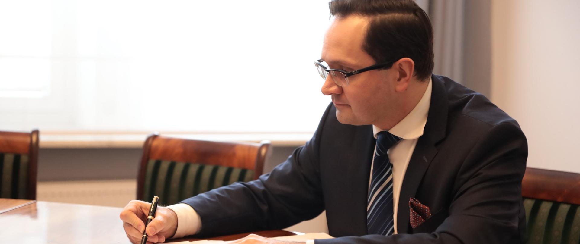 Wiceminister Mariusz Golecki podpisuje dokumenty. Siedzi przy stole i w prawym ręku trzyma pióro.