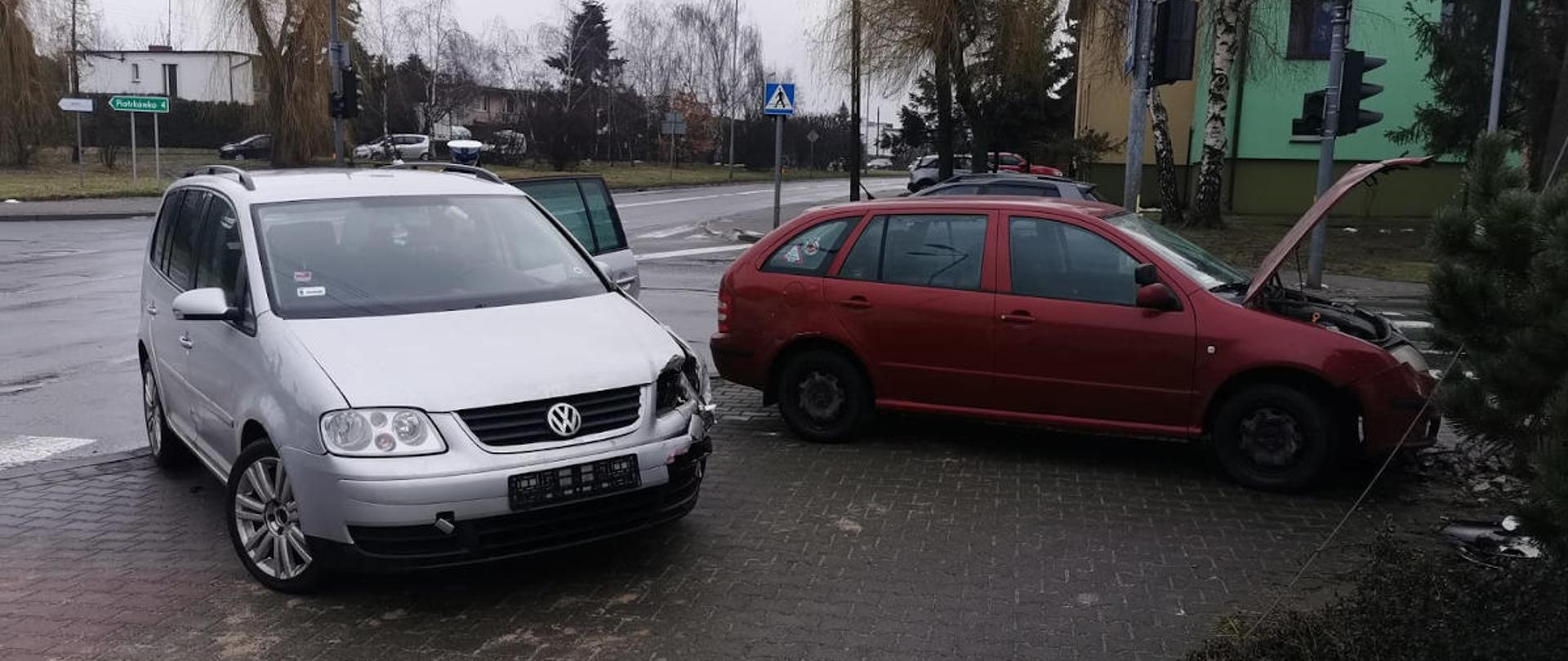 Widać dwa uszkodzone samochody na poboczu, znajdują się blisko siebie, w oddali kilka drzew