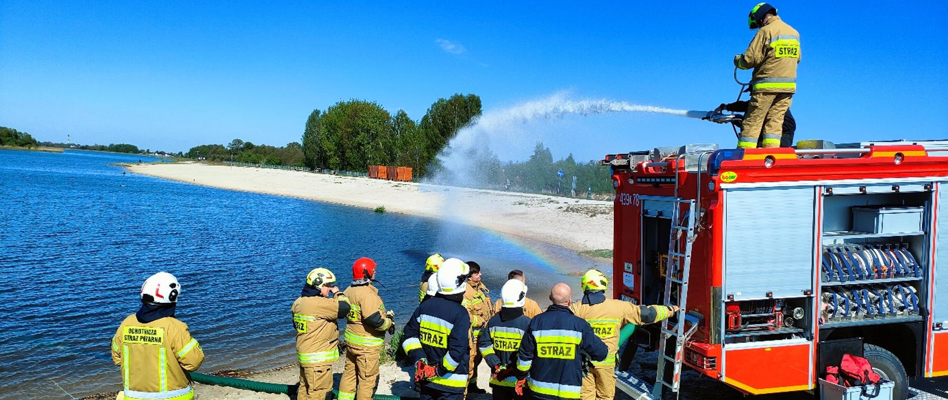 Zdjęcie przedstawia strażaków podczas ćwiczeń z poboru wody z naturalnego zbiornika, najprawdopodobniej rzeki lub jeziora. Grupa strażaków z Państwowej i Ochotniczej Straży Pożarnej, ubranych w beżowe i czarne mundury ochronne z odblaskowymi oznaczeniami, bierze udział w operacji. Część z nich obsługuje węże ssawne, które pobierają wodę z rzeki, a inni stoją przy czerwonym wozie strażackim z otwartymi schowkami na sprzęt.
Na dachu pojazdu znajduje się strażak obsługujący działko wodne, które wyrzuca strumień wody na dużą odległość. W wyniku rozpylania wody powstała tęcza, dodająca dynamicznego efektu wizualnego.
Teren ćwiczeń obejmuje piaszczystą plażę nad wodą, z zielenią i drzewami w tle. W oddali widoczne są pomarańczowe kontenery i znaki drogowe, co sugeruje, że miejsce to może być przeznaczone do rekreacji lub ćwiczeń ratowniczych. Niebo jest bezchmurne, a słońce jasno oświetla całą scenę, co wskazuje na sprzyjające warunki pogodowe.