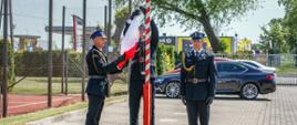 Na zdjęciu moment zawieszenia flagi państwowej na maszt przez strażaków w mundurach wyjściowych koloru czarnego na tle zaparkowanych pojazdów.
