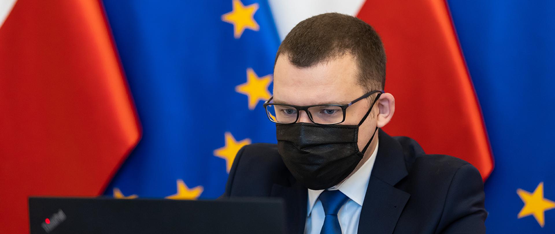 Na zdjęcie widać wiceministra Pawła Szefernakera patrzącego w ekran na tle flag Polski i UE.