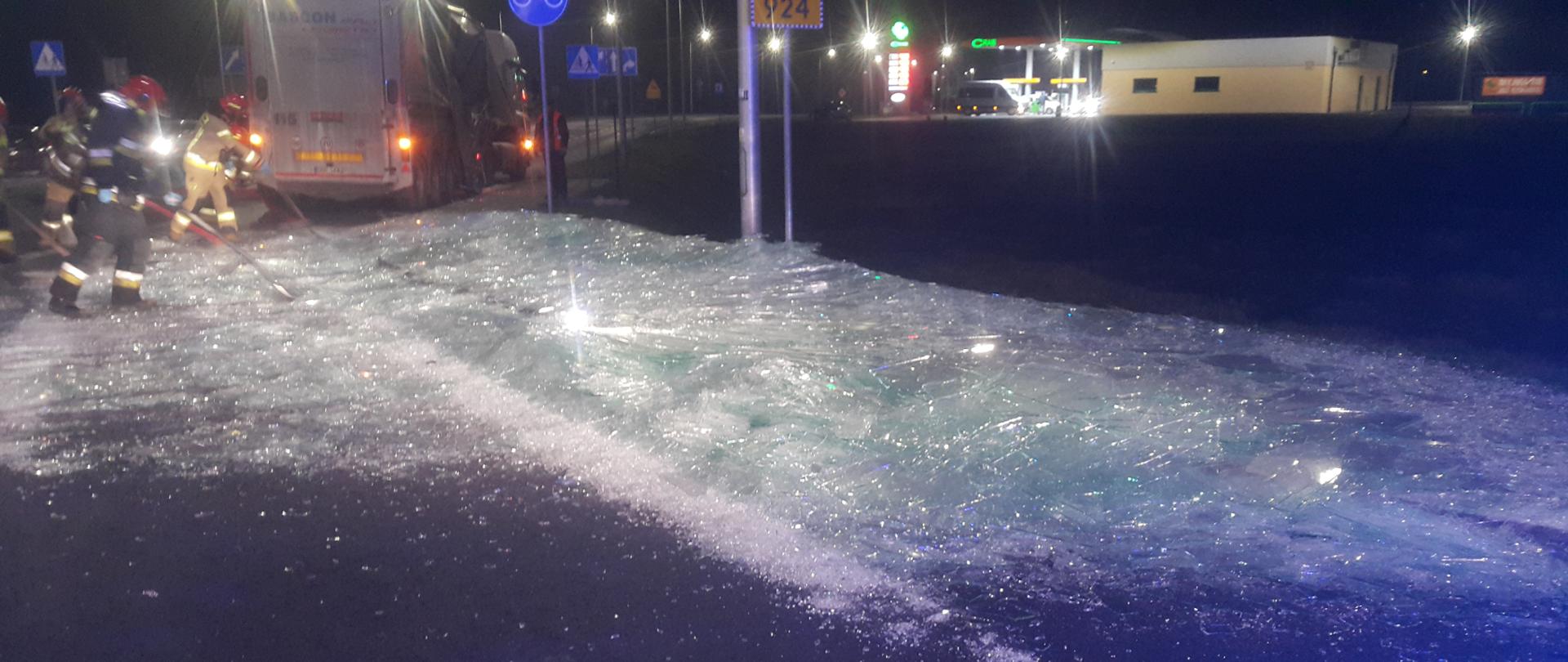 Rozsypane potłuczone szkło na jezdni, w dalszej części zdjęcia strażacy zgarniający szkło na pobocze, samochód ciężarowy oraz stacja paliw.