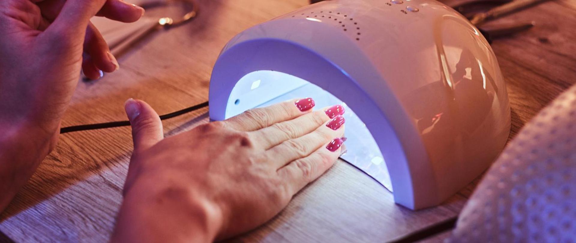Dłoń z pomalowanymi na różowo paznokciami umieszczona pod lampą UV/LED do utwardzania lakieru hybrydowego w salonie kosmetycznym.