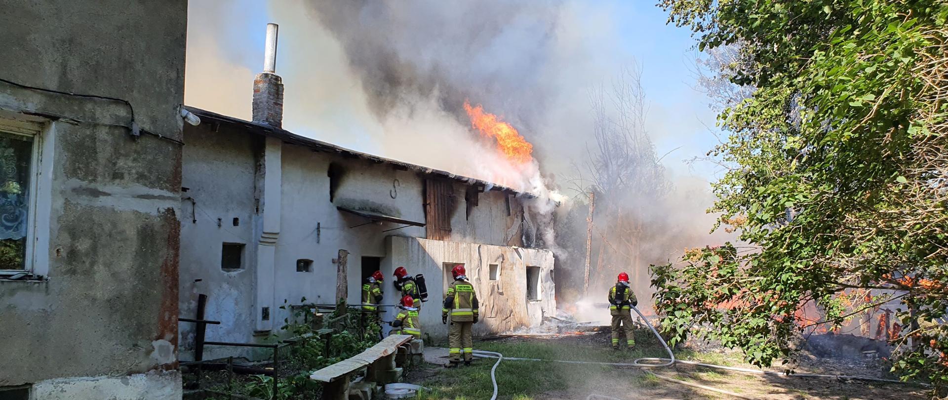 Zdjęcie przedstawia kompleks budynków murowanych, na końcu którego widoczne są płomienie na dachu oraz chmura dymu. Przed budynkiem znajduje się 5 strażaków, którzy gaszą pożar.