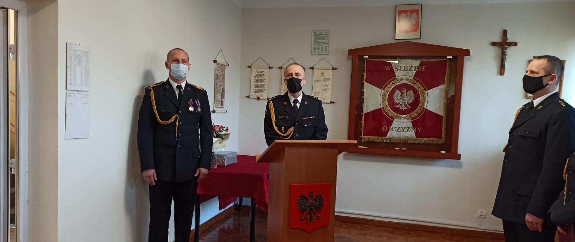 Zdjęcie przedstawia 3 mężczyzn. Obok nich znajduje się sztandar KP PSP Świdwin, mównica przed którą przemawia komendant powiatowy, Godło Rzeczypospolitej Polskiej oraz Krzyż.