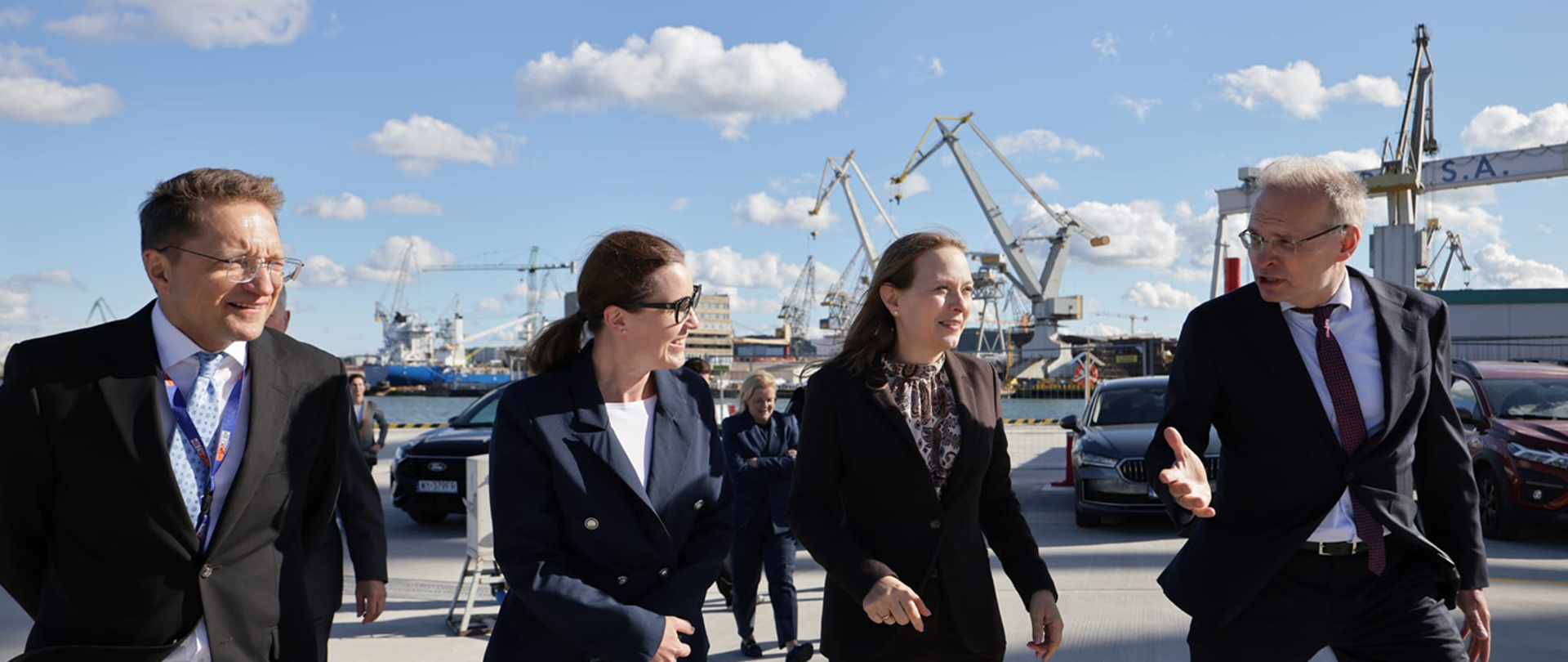 Minister funduszy i polityki regionalnej Katarzyna Pełczyńska-Nałęcz idzie z przedstawicielami portu i rozmawia z jednym z nich. W tle znajdują się portowe dźwigi i zacumowane jednostki.
